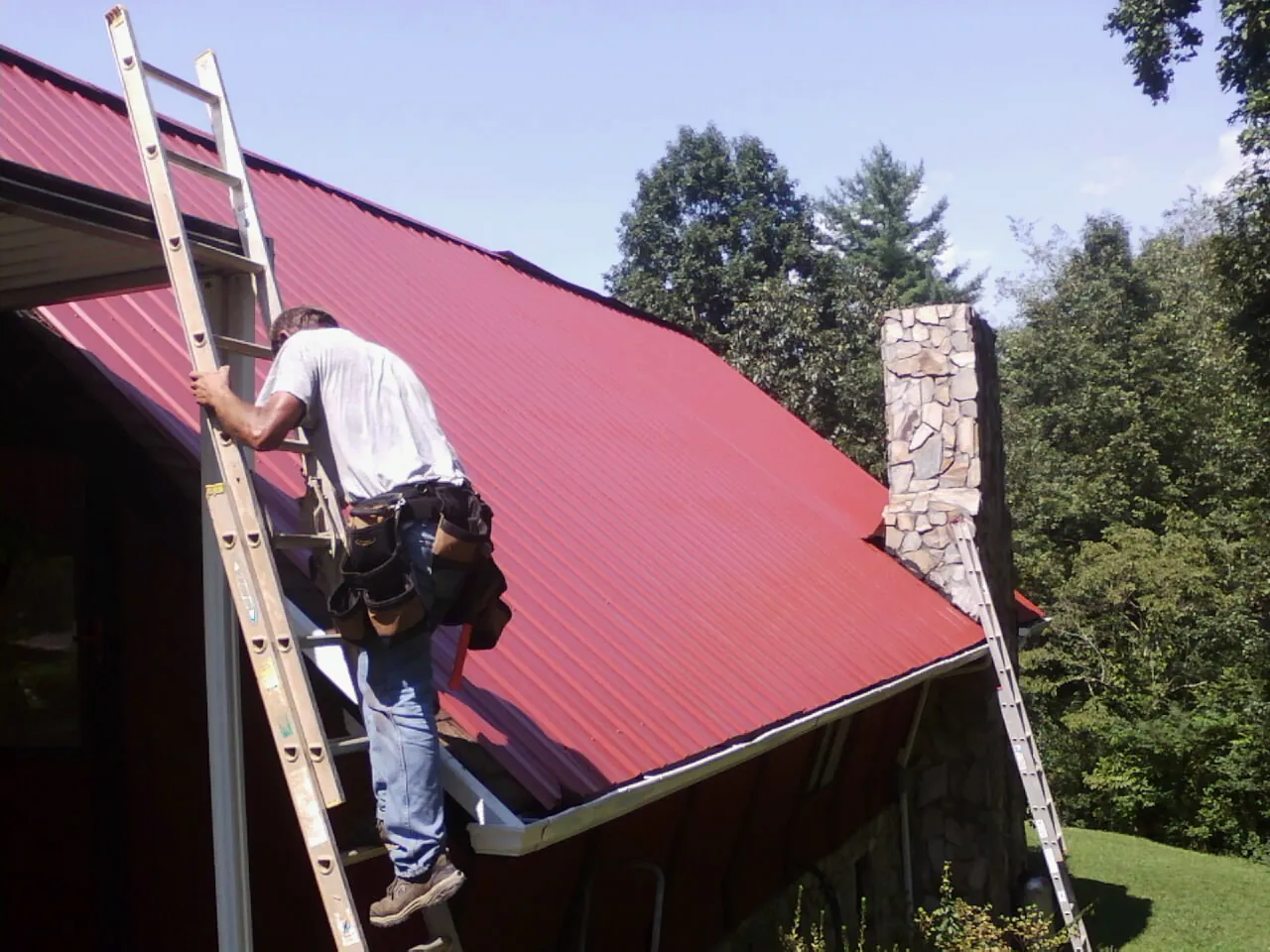 Roofer on a ladder beside a red metal roof and stone chimney — Soffit & Fascia Repair by Roofing Brothers in Mojave